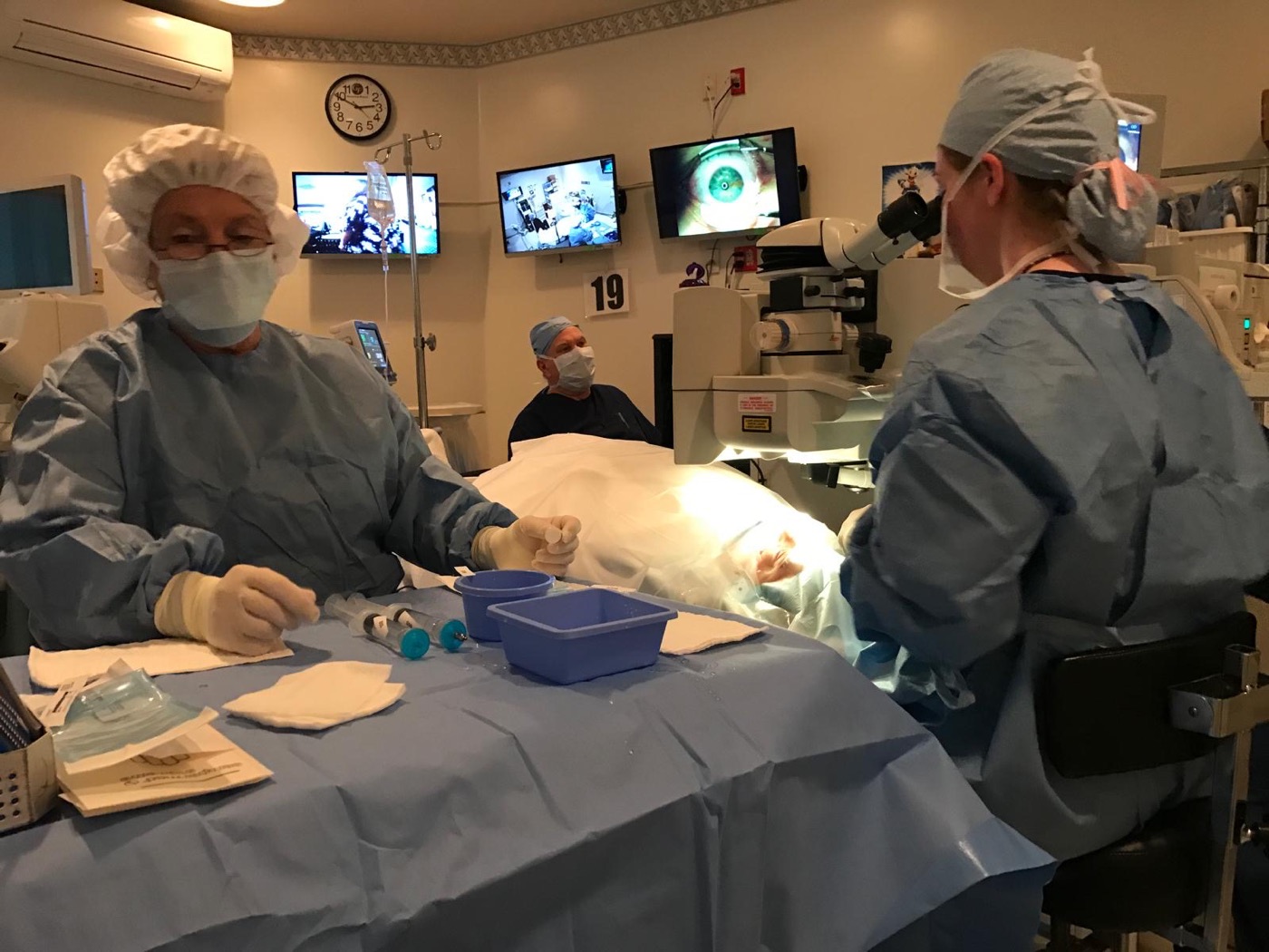 Two surgical staff at the operating microscope, the eye magnified on the monitors behind them.