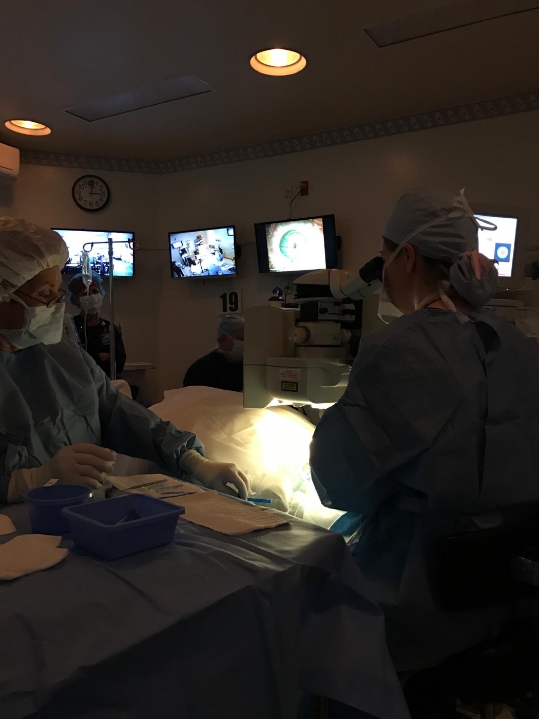 The operating room mid-procedure, with the surgical field and the magnified eye displayed across the monitors.